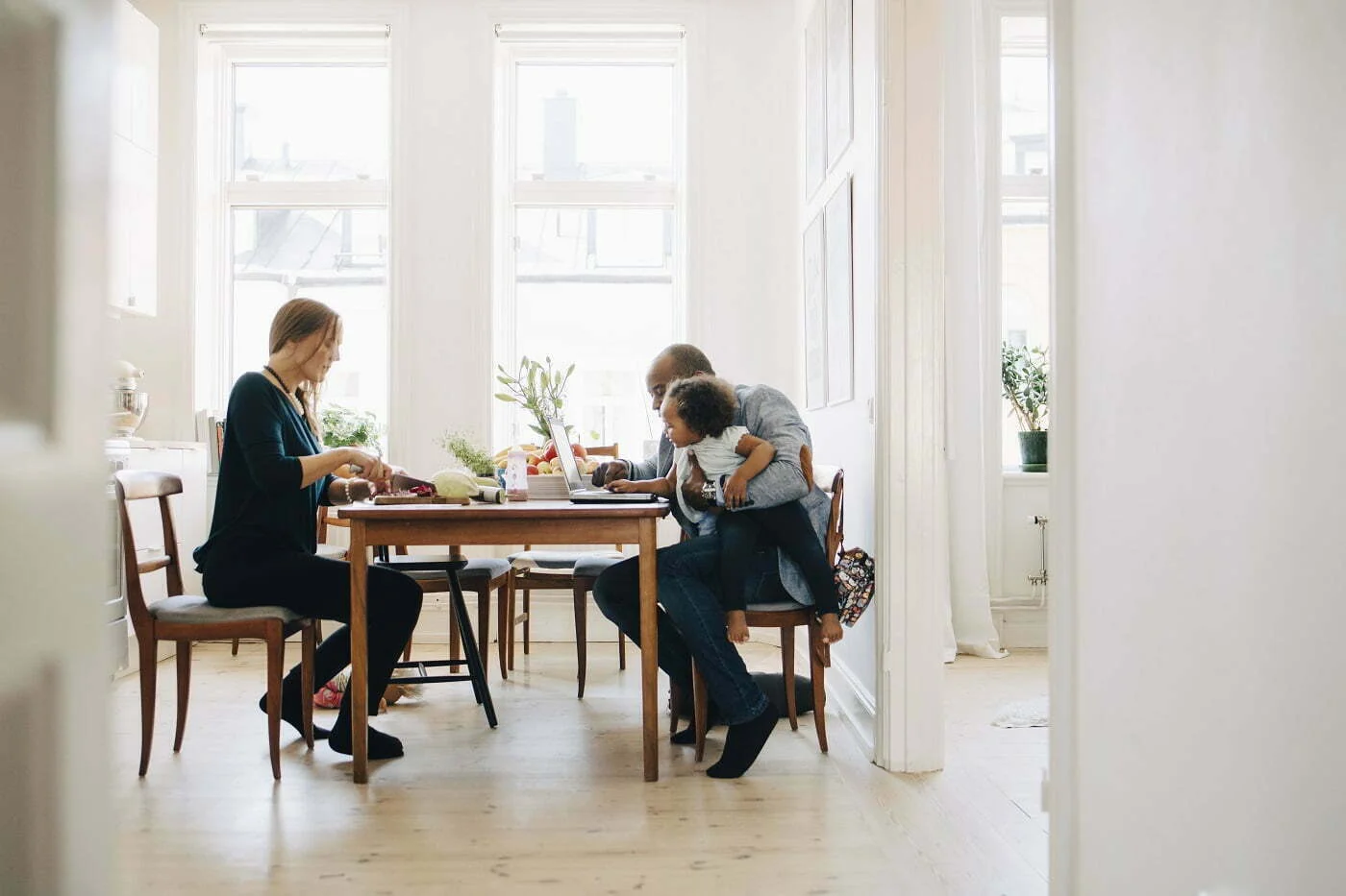 Äiti, isä ja taapero viettävät yhdessä vapaa aikaansa pöydän äärellä. Äiti tekee ruokaa ja isä selaa tablettia taapero sylissään Retta
