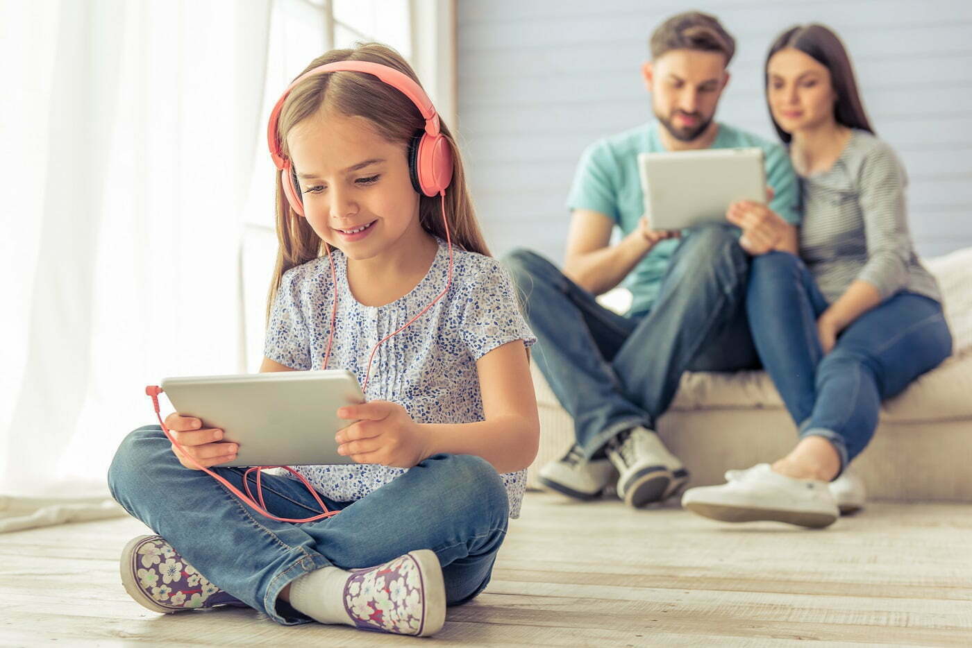 Pieni tyttö katsoo tablettia kuulokkeet päässä ja hymyilee lattialla istuen. Vanhemmat istuvat tytön takana Retta